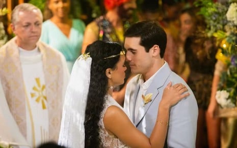 Luna usa um vestido de noiva e véu e abraça Nicolas Prattes, que está com um terno cinza claro; os dois estão prestes a se beijar
