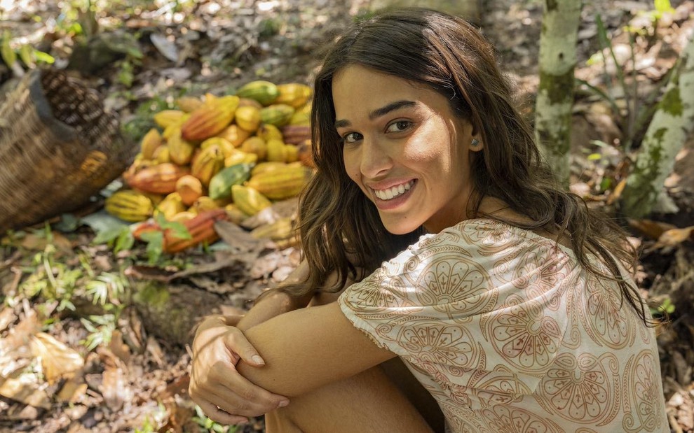 A atriz Thereza Fonseca está sentada em um jardim caracterizada como Mariana no remake de Renascer, da Globo