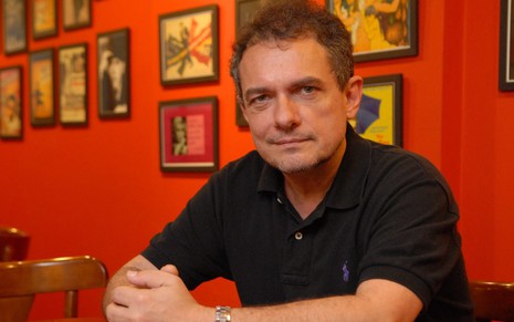 O diretor e roteirista João Falcão com expressão séria, olhando para a câmera, com parede vermelha ao fundo