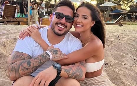 João Hadad e Luana Andrade abraçados, numa praia, sorrindo para foto