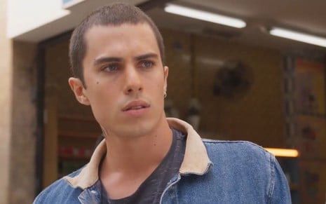 O ator Luan Argollo usa jaqueta jeans e exibe expressão séria em cena como Marcos na novela Elas por Elas