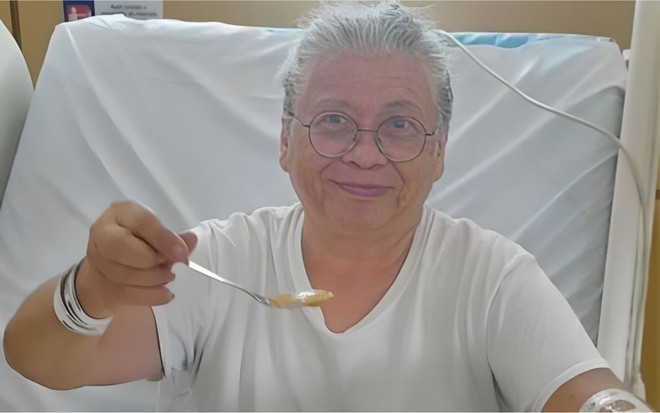 Marlene Mattos com cabelo grisalho preso, segurando uma colher e sorrindo, deitada em uma cama hospitalar