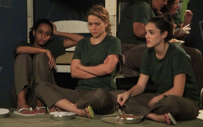 As atrizes Taís Araujo, Leandra Leal e Isabelle Drummond com expressões tristes e uniformes de presidiárias, sentadas no chão, com quentinhas à frente, em cena de Cheias de Charme