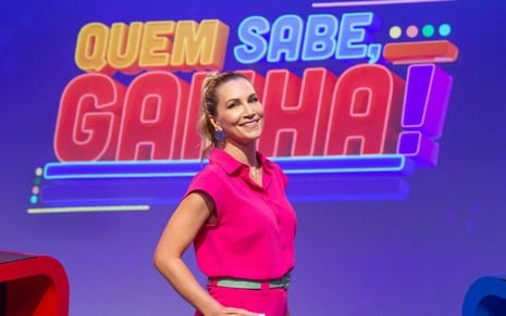 A apresentadora Anne Lottermann sorrinfo, de macacão rosa pink, no estúdio do game show que apresentará, Quem Sabe, Ganha!