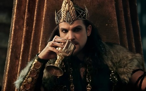 Igor Rickli está vestido como rei, com coroa na cabeça e trajes elegantes; ele bebe uma taça de vinho
