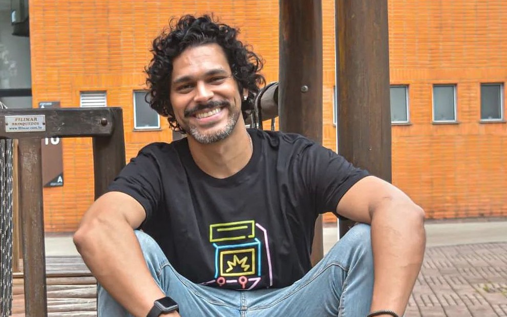 De camiseta azul e calça jeans, Renan Monteiro está sentado com os braços apoiados na perna
