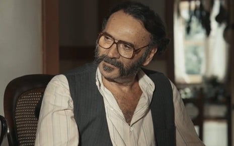 O ator Almir Sater com expressão séria em cena como Rachid em Renascer, de óculos, camisa branca aberta no peito, colete preto