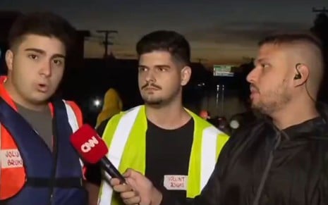 Um homem com um colete vermelho grita ao lado de um homem com colete verde ao lado do repórter Pedro Teixeira na CNN Brasil
