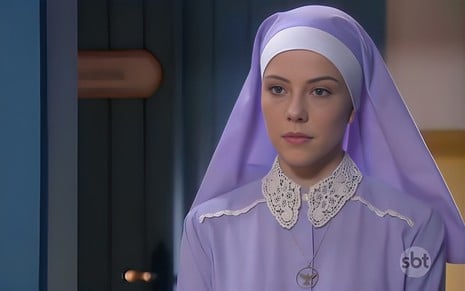 Bia Arantes está vestida como freira, com hábito lilás, e tem expressão séria em cena de Carinha de Anjo