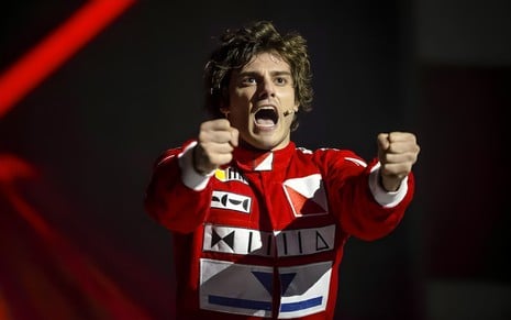 Vestido com macacão vermelho de piloto de Fórmula 1, Hugo Bonemer segura um volante imaginário e grita