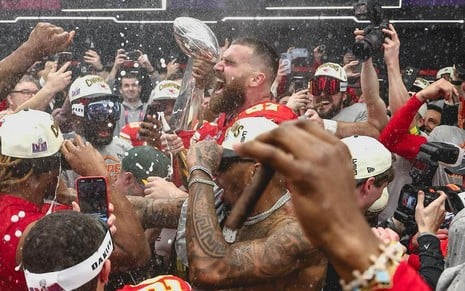 Travis Kelce grita com o troféu do Super Bowl ao fundo e em meio ao time do Chiefs; há chuva de champanhe para todo lado