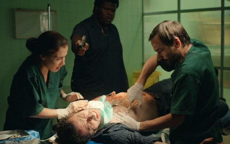 Evandro (Julio Andrade) e Carolina (Marjorie Estiano) operam Aristeu (Cesar Ferrario) sob mira de revólver - Reprodução/TV Globo