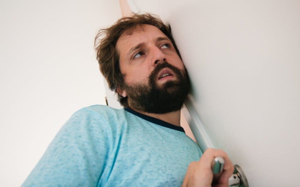 O ator Gregório Duvivier com ouvido colado numa porta, de camiseta azul e expressão séria, em cena da série 5X Comédia