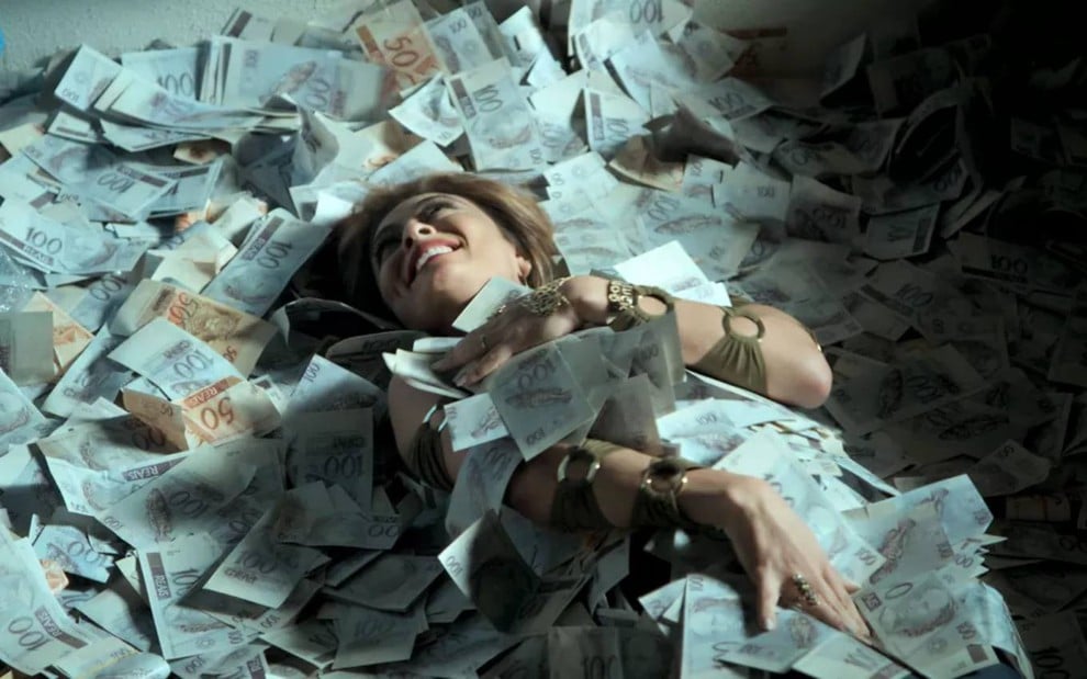 A atriz Juliana Paes deitada em cima de muitas notas de dinheiro e sorrindo em cena de A Força do Querer