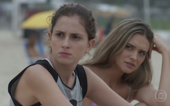 A atriz Carol Duarte com expressão emburrada e Juliana Paiva atrás, também de cara fechada, em cena de A Força do Querer
