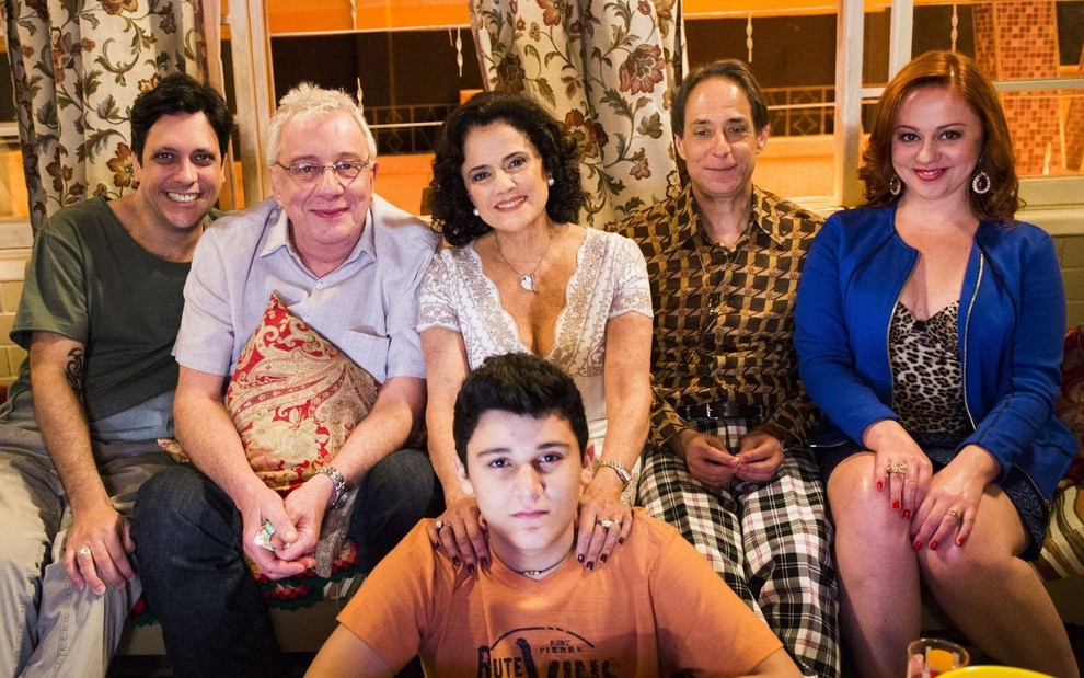 Atores de A Grande Família sorriem para foto sentados em sofá da casa cenográfica da série