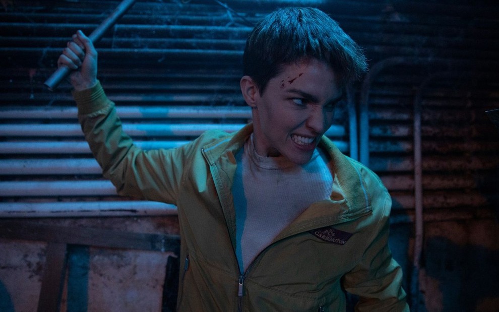 Ruby Rose com os cabelos bem curtos em cena do filme A Protetora (2020); ela pega um pedaço de pau e ameaça bater em alguém