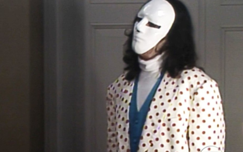 O ator Breno Moroni em cena como o Mascarado de A Viagem; ele usa máscara branca cobrindo todo o rosto e terno branco com bolinhas vermelhas