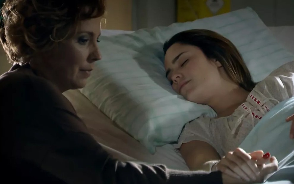 A atriz Ana Beatriz Nogueira ao lado de Fernanda Vasconcellos, em cena em cama de hospital; Fernanda dormindo e Ana Beatriz segurando a mão dela