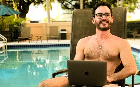 Aaron Salles Torres sem camisa, com laptop no colo, sentado à beira de uma piscina