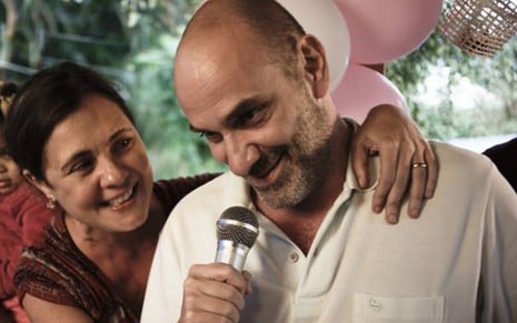 Adriana Esteves abraçada com o Marco Ricca, sorrindo enquanto segura um microfone perto da boca dele