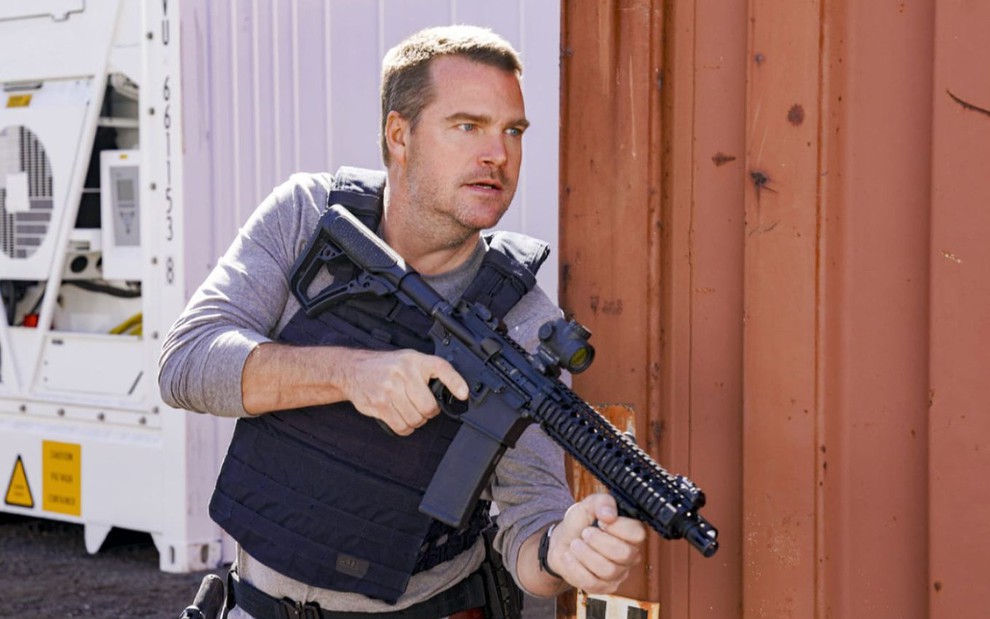 Chris O'Donnell segura uma arma e se esconde atrás de um contêiner em cena de NCIS: Los Angeles