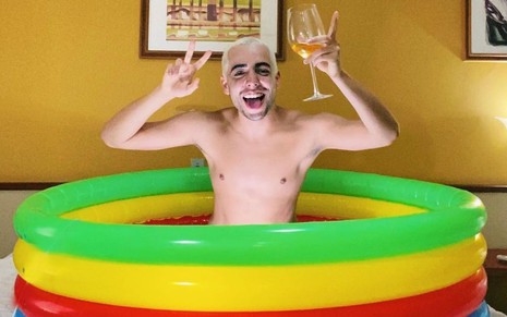 Imagem de Lucas Selfie, sem camisa, dentro de uma piscina inflável, segurando uma taça de bebida