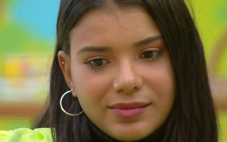 Jakelyne Oliveira está olhando para baixo, usa um colete verde e está de cabelo solto; ela está na frente de um fundo colorido