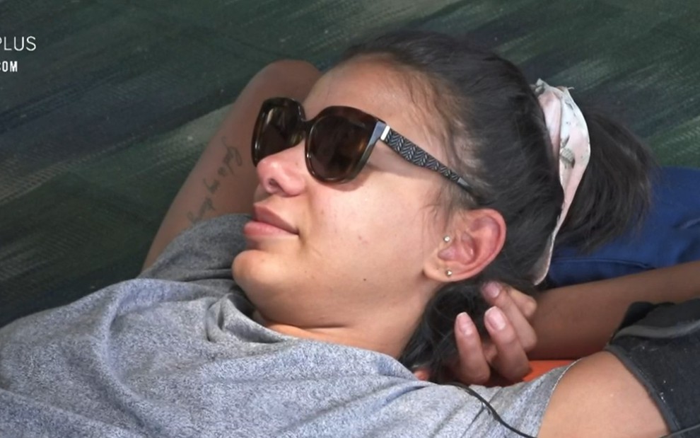 Jakelyne está deitada no chão na casa da árvore; a peoa usa lenço branco no cabelo preso; ela está de óculos e camiseta cinza