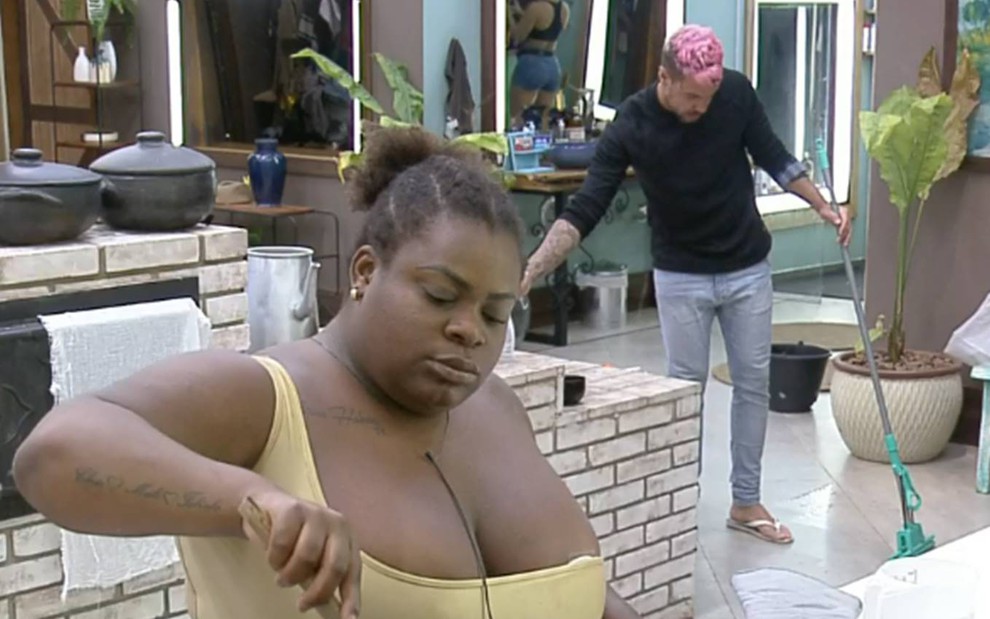 Jojo Todynho usa blusa de alça amarela, enquanto prepara almoço na cozinha da sede em A Fazenda, ao fundo, Lipe Ribeiro usa blusa preta, calça jeans, está de cabelo rosa, enquanto varre o chão da cozinha