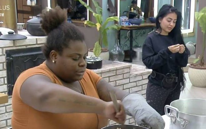 Jojo está na cozinha da sede de A Fazenda 12, preparando o almoço, ela usa uma camiseta laranja e está de cabelo preso. Ao lado dela, está MC Mirella com um moletom preto e calça preta. A funkeira está de cabelo solto