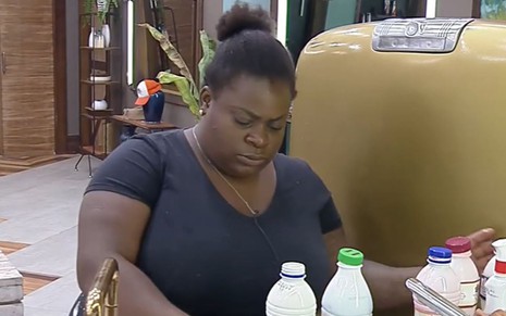 Jojo está na cozinha da sede do reality; a cantora usa uma camiseta preta e está de cabelo preso; ela olha para baixo e separa embalagens de leite e outros alimentos ao lado da geladeira