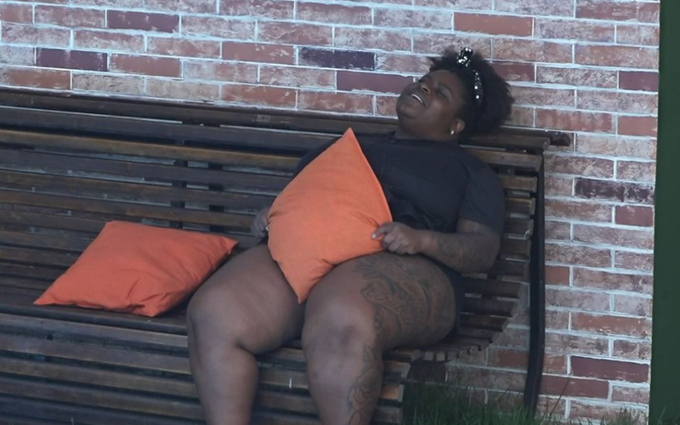Jojo está sentada no banco na área externa da sede; peoa usa roupa preta e está segurando uma almofada laranja