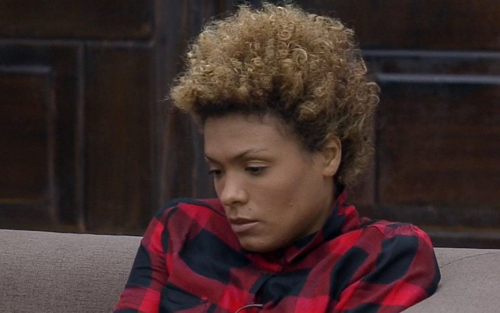 Lidi Lisboa está sentada no sofá da sede; ela usa uma blusa xadrez vermelha com detalhes pretos; a atriz olha para baixo e parece pensativa