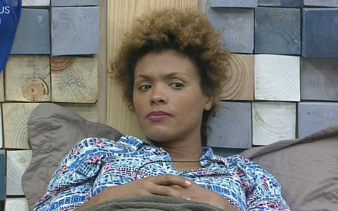 Lidi Lisboa está com camiseta azul com detalhes coloridos; olha para o lado; está encostada na parede