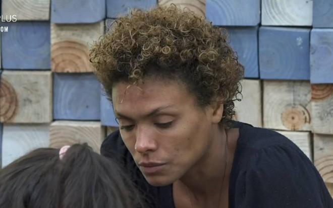 Lidi aparece olhando para baixo; ela está usando uma blusa preta e o fundo é azul