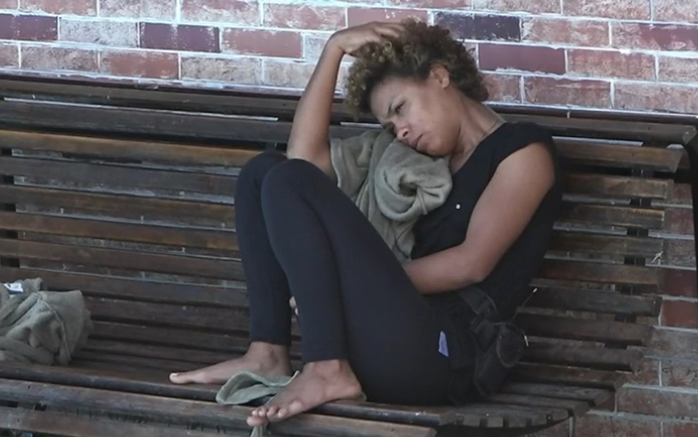 Lidi está sentada em um banco na área externa da sede; a peoa está encostada no ombro, com a mão na cabeça; ela usa camiseta preta e calça preta