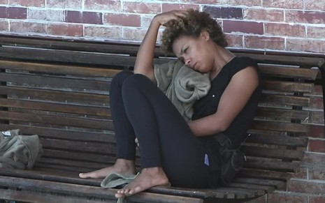Lidi está sentada em um banco na área externa da sede; a peoa está encostada no ombro, com a mão na cabeça; ela usa camiseta preta e calça preta
