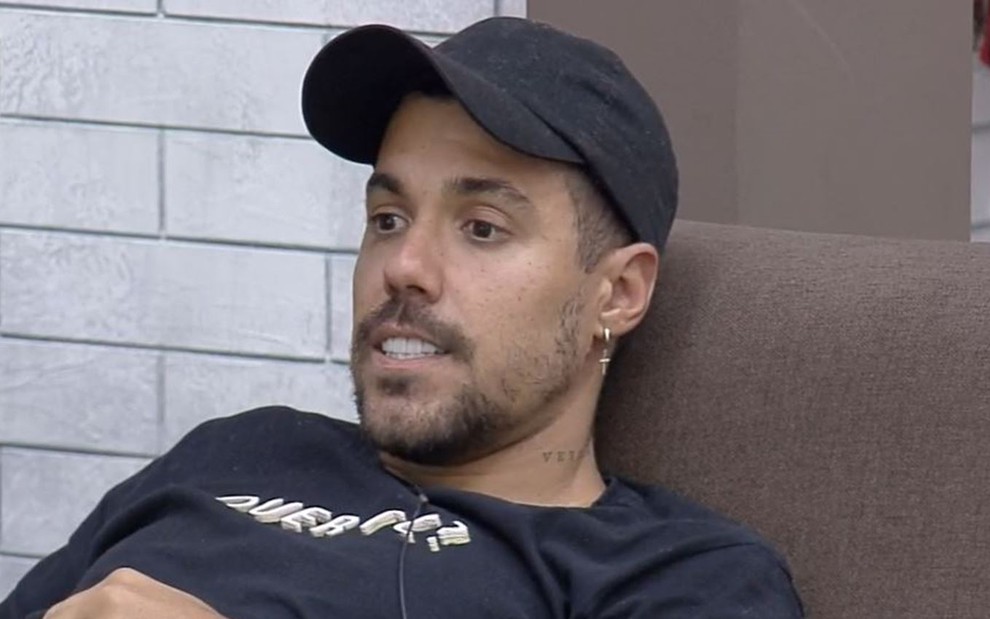 Lipe Ribeiro está sentado no sofá da sede; ele usa boné preto, camiseta preta e olha para longe