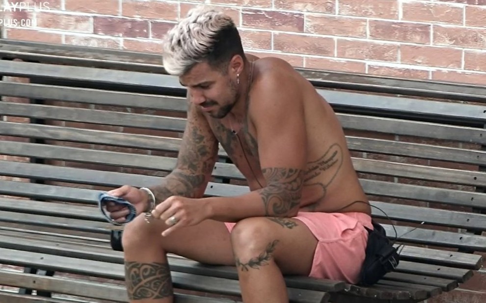 Lipe Ribeiro está sentado no banco da área externa da sede; o peão está sem camiseta e usa bermuda rosa; ele olha para baixo