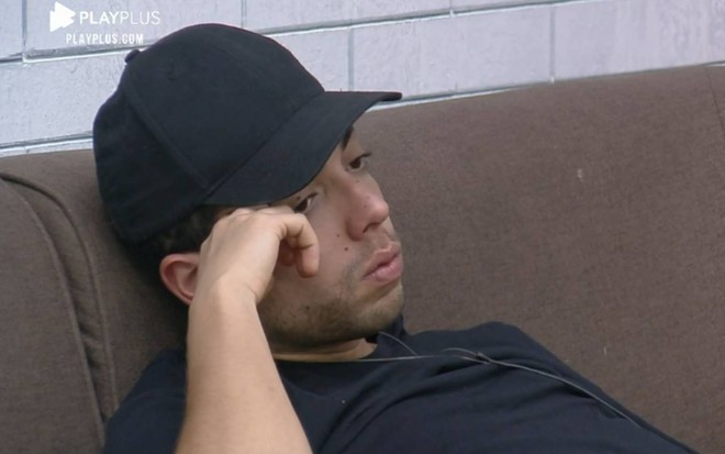 Lucas está no sofá da sede de A Fazenda 12; o peão usa um boné preto, camiseta preta e está com a mão no rosto, encostado no sofá