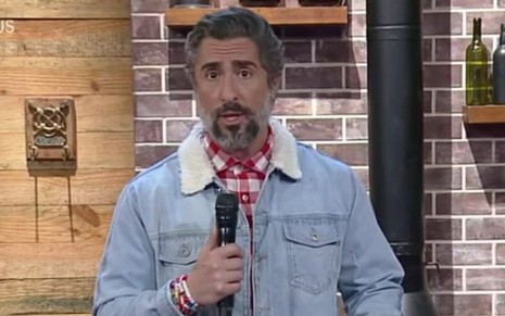 Marcos Mion aparece apresentando o programa ao vivo; ele usa jaqueta azul clara; está com camiseta xadrez vermelha e branca por baixo; ele segura o microfone e encara a câmera