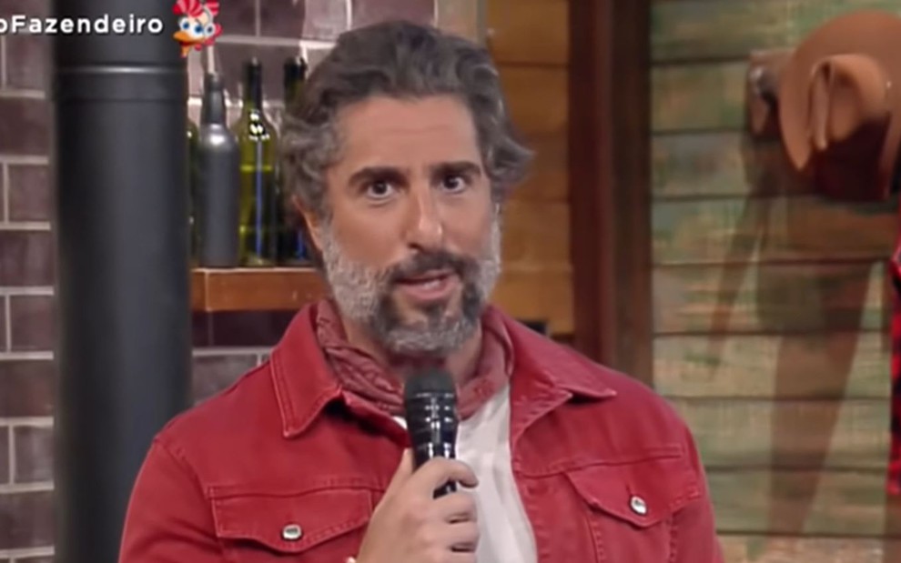 Marcos Mion aparece no estúdio de A Fazenda 12, ele usa camiseta branca, lenço no pescoço vermelho e jaqueta vermelha; o apresentador segura o microfone