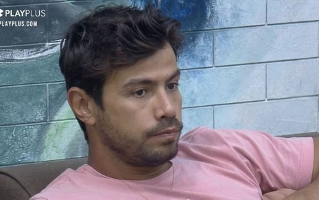 Mariano está sentado no sofá e olha para frente; o peão está com camiseta rosa; ele está na frente de uma parede azul