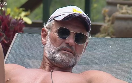 Mateus Carrieri está tomando sol na área da piscina; ator usa boné branco e óculos escuro