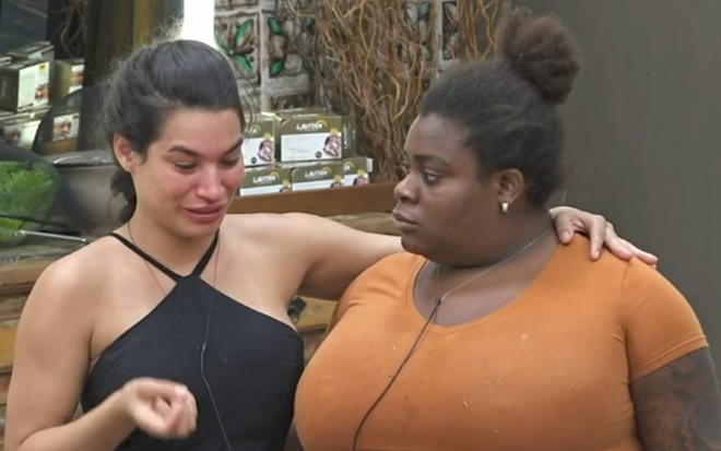Raissa está com uma blusa preta, com alça trançada e aparece chorando ao lado de Jojo. A funkeira abraça a amiga; ela usa cabelo preso e camiseta laranja