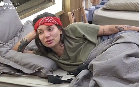 Stéfani está deitada na cama; a peoa usa uma bandana vermelha e está de cabelo solto; ela está vestindo uma camiseta verde militar