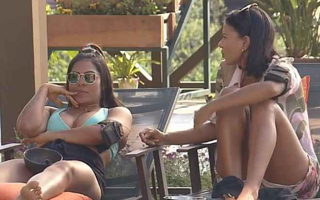 Tays está sentada ao lado de Jakelyne na área da piscina, usa um top azul e óculos escuro. Jakelyne está virada de lado com uma camiseta com tons rosa e cinza