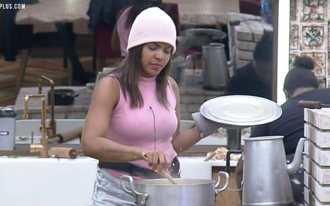 Tays está na cozinha da sede; ela mexe a comida que está na panela e segura a tampa com a outra mão; ela usa uma blusa rosa e touca cor de rosa; a peoa está de cabelo solto