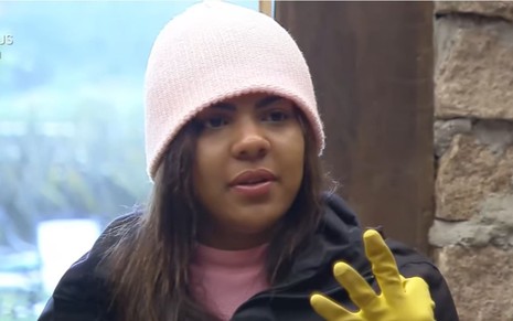Tays Reis está usando uma jaqueta preta e uma blusa rosa; a peoa usa ainda uma luva amarela e touca cor de rosa; ela faz um sinal com os três dedos da mão e olha olha frente